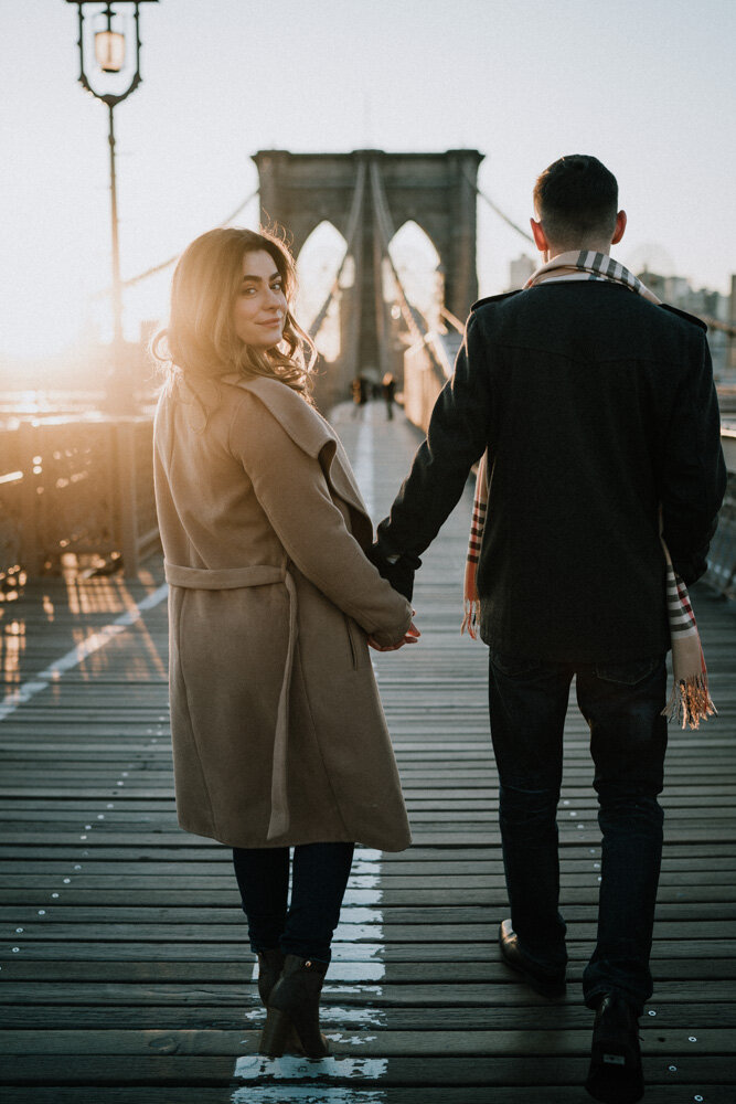 Woman holds boyfriend's hand on brooklyn bridge and glances back at photographer