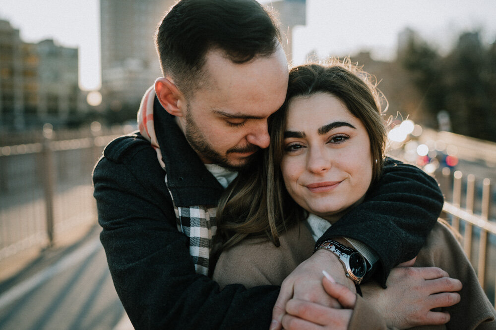 Man wraps arms around girlfriend's shoulders while she looks at photographer