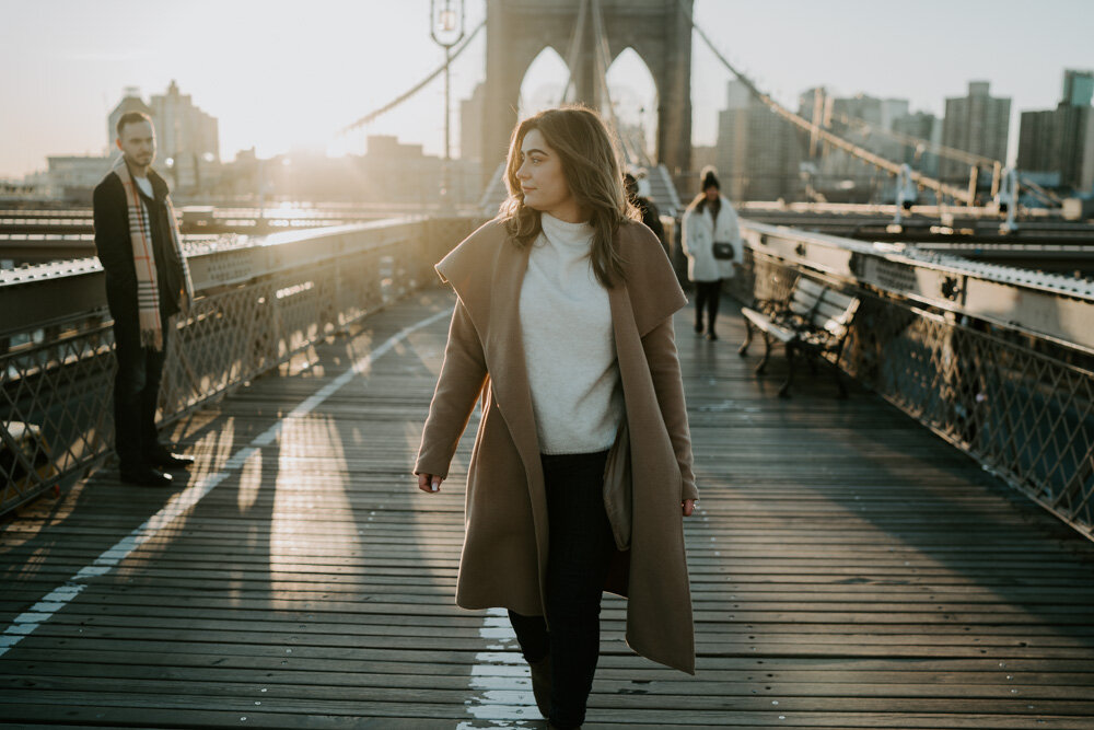 Woman walks on brooklyn bridge at sunrise looking at her fiancé