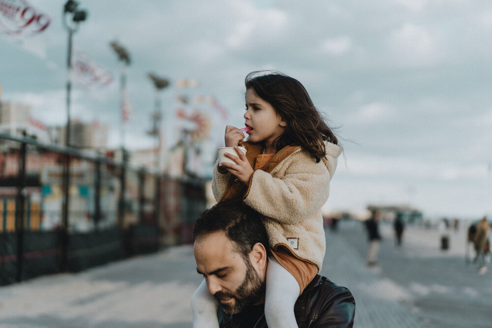 Young girl eats ice cream on dad's shoulders during family session at coney island
