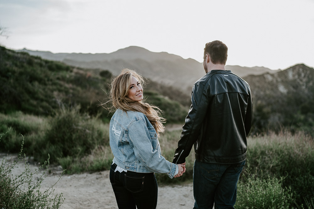 Woman holds boyfriend's hand outdoors and glances back at photographer